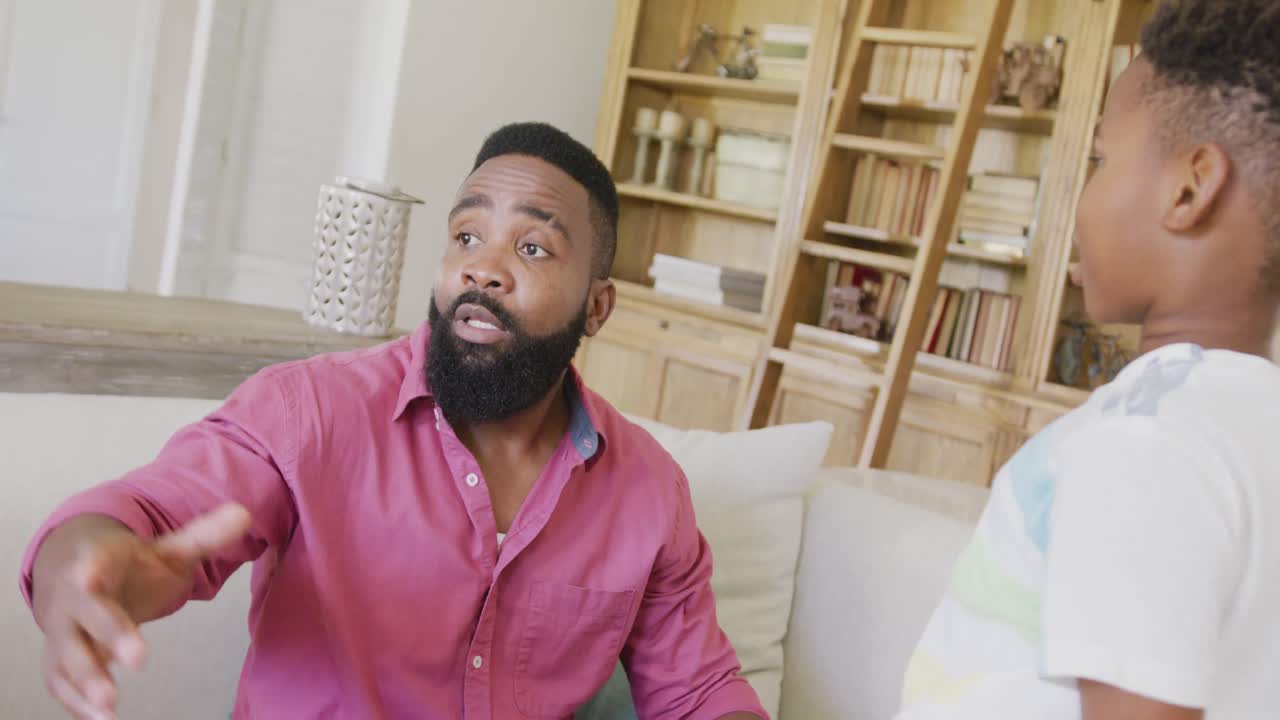 Serious african american father and son sitting on sofa and talking, in slow motion