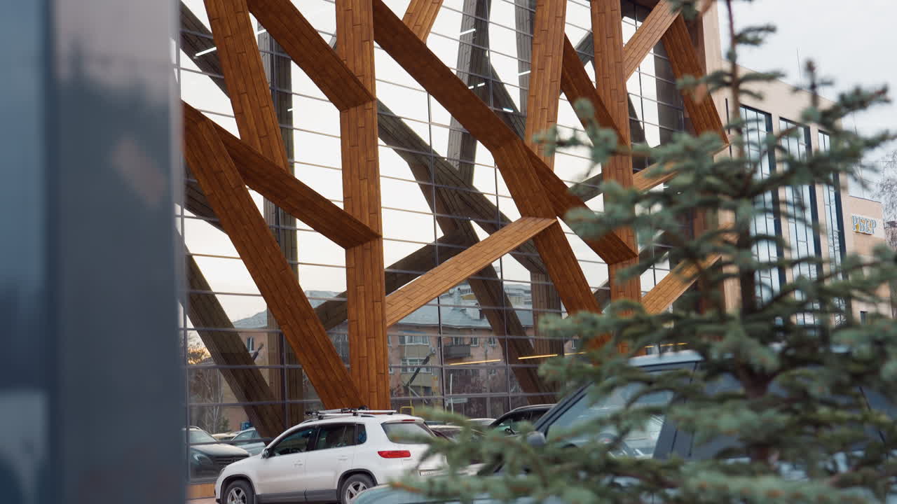 Modern gym exterior shot from upper angle showing geometric wooden beams and floor-to-ceiling glass panels, surrounding parking area with cars and evergreen trees under overcast sky