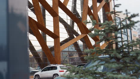 modern gym exterior shot from upper angle showing geometric wooden beams and floor-to-ceiling glass panels, surrounding parking area with cars and evergreen trees under overcast sky