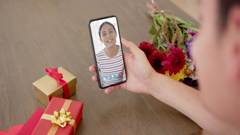 mujer biracial sosteniendo un teléfono inteligente con una mujer hablando en la pantalla con regalos en el escritorio