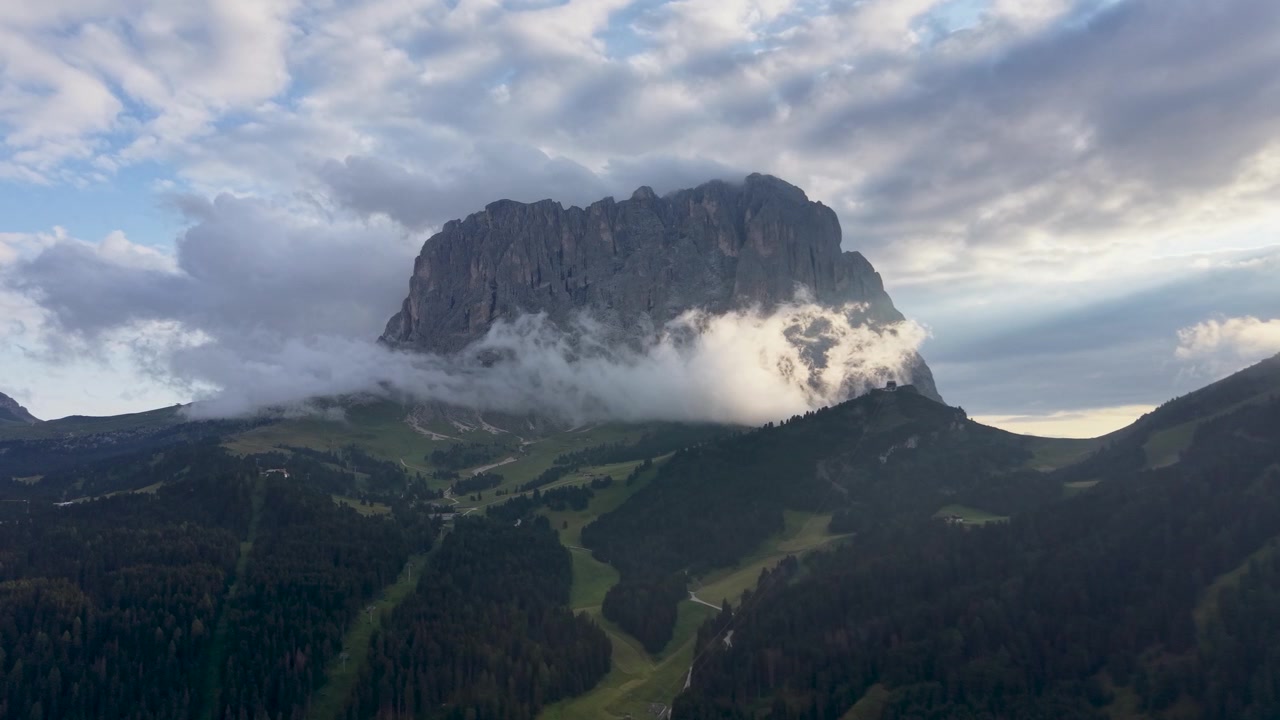 Vista aerea con drone del Gruppo del Sella che si erge sopra pendii boscosi e prati alpini, con nuvole alla deriva che circondano il massiccio calcareo aspro nelle Dolomiti.
