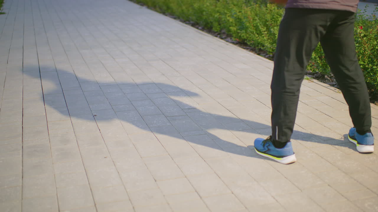 park sidewalk shadow of person standing in athletic stance on tiled pavement, blue sneakers visible with casual pants, long cast shadow stretching along path, green hedge border, sunny afternoon