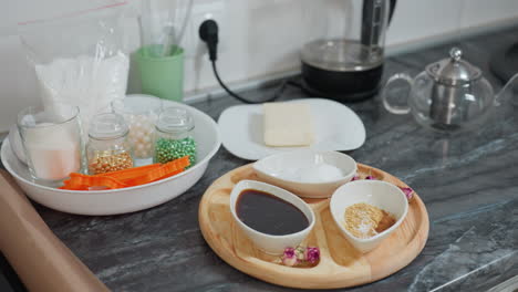 close-up of modern kitchen countertop featuring wooden tray with ceramic bowls containing sauce, sugar, and spices, butter on plate, glass jars with ingredients, and glass kettle on black marble