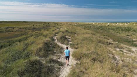 aerial follow of male walking on sand path amongs sand dunes