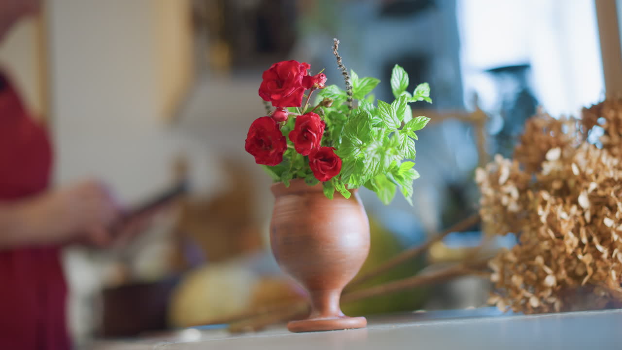 Jarrón en la encimera de la cocina, flores rojas, cocinero casero difuminado al fondo, luz cálida de la mañana, jarrón de cerámica rústico, follaje seco, bokeh suave, atmósfera doméstica íntima, sutil movimiento de manos.