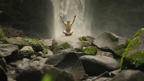 fit woman in white bikini raising arms in easy pose on rock at powerful waterfall