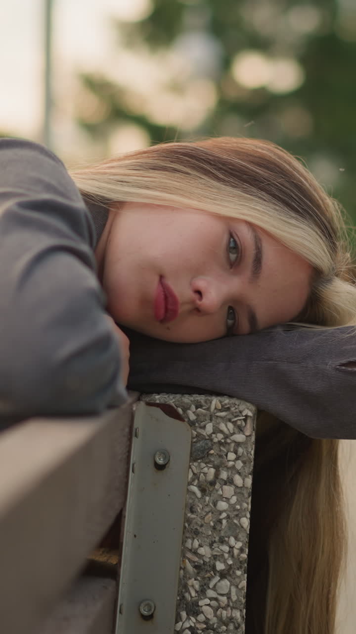 Woman in grey clothing resting her head on arm outdoor, lost in thought with a peaceful expression, the background is blurred, highlighting her thoughtful state