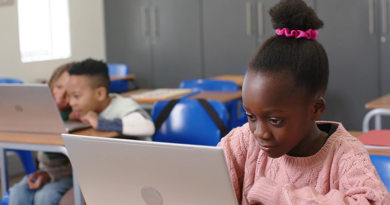 Bambino afroamericano con maglione rosa che aggrotta la fronte, si appoggia e digita su un laptop argentato alla scrivania, con spazio per il testo