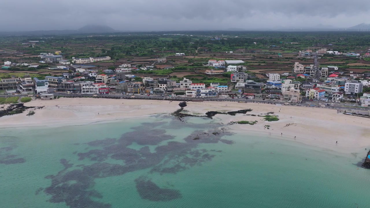 Güney Kore'nin Jeju Adası'ndaki Gimnyeong Plajı boyunca sağa doğru hareket eden yatay bir çekim; turkuaz suyu, kumlu plajı, köyü, kıyı şeridini, tarım arazilerini ve bulutlu gökyüzünü gösteriyor.