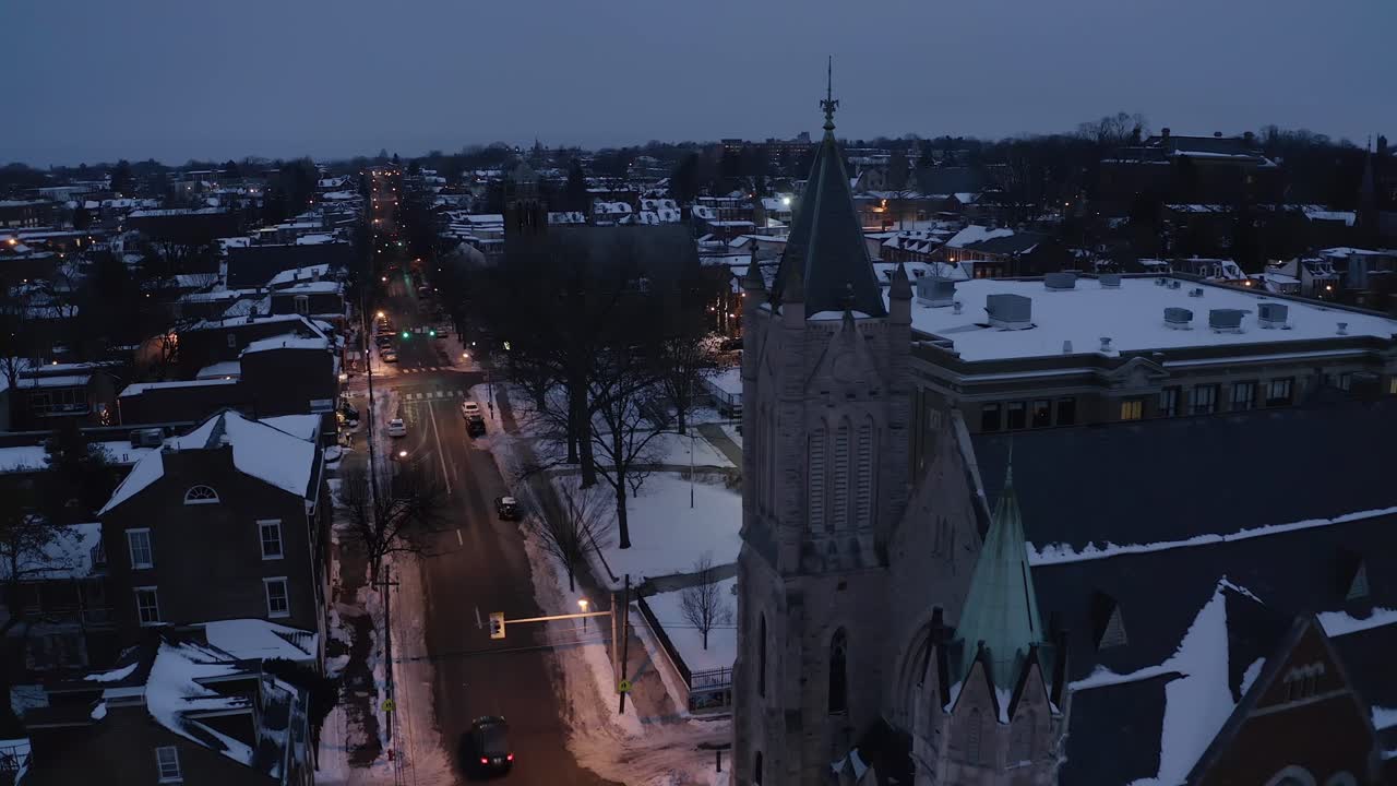 antena de una calle tranquila de la ciudad durante la nieve invernal mientras el dron pasa por la iglesia y la escuela al atardecer