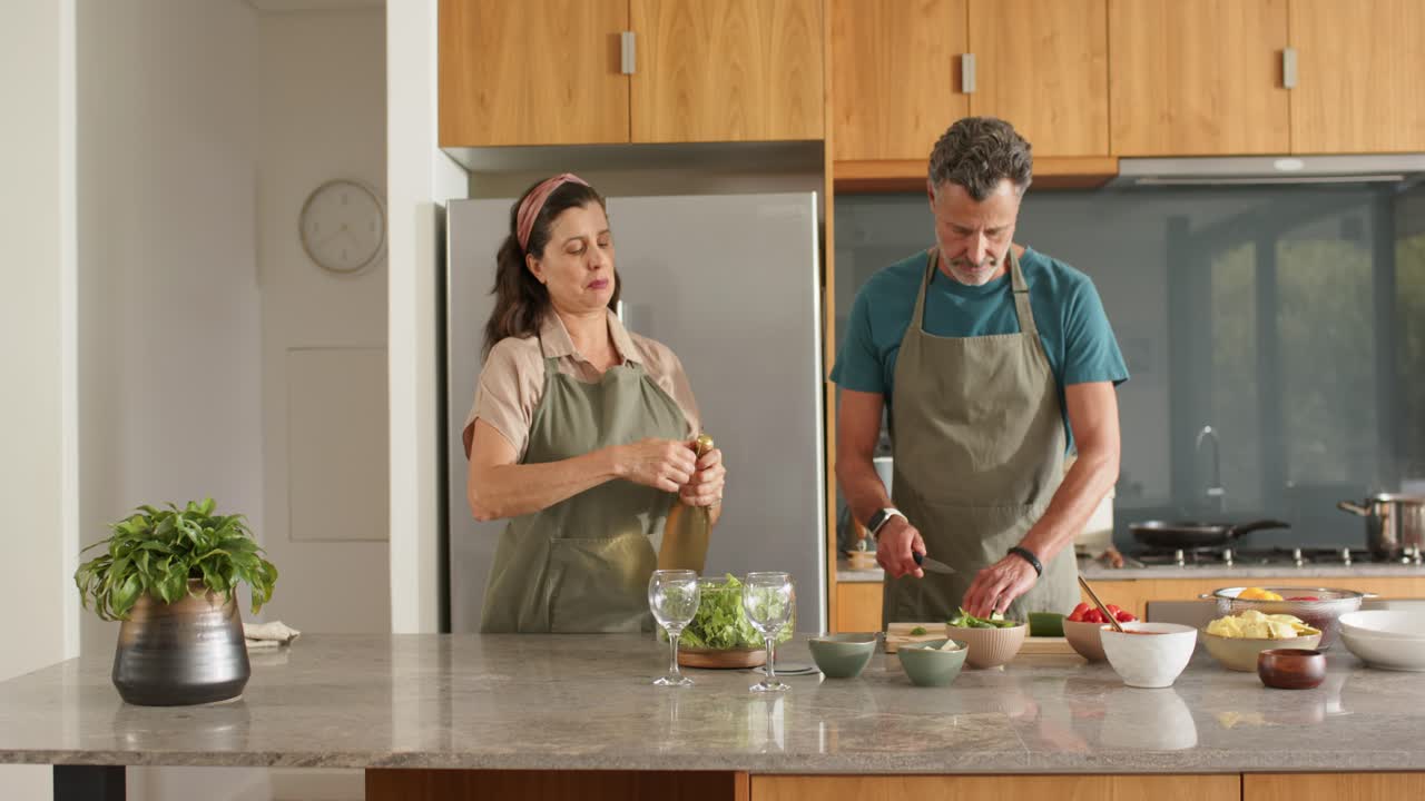 Una coppia in grembiule cucina in cucina, la donna apre una bottiglia sigillata con un apribottiglie mentre l'uomo taglia le verdure.