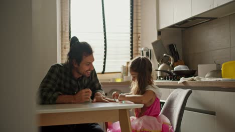 Shooting-from-behind-the-wall,-a-happy-man-in-a-green-checkered-shirt-is-sitting-at-the-kitchen-table-with-his-little-daughter,-a-brunette-girl-in-a-pink-dress,-who-is-doing-a-manicure-for-her-father-and-they-are-playing-together-in-the-kitchen