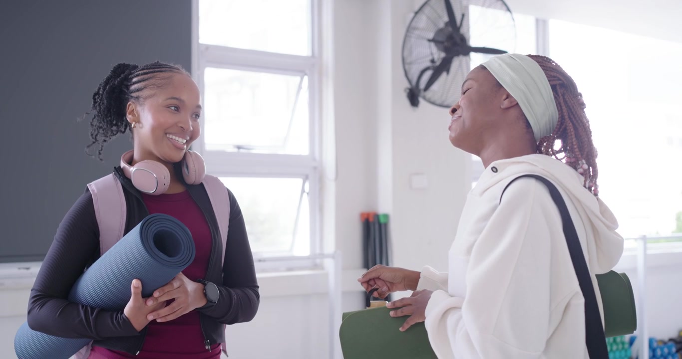 Two African women meeting before class in studio, holding mats and chatting about workout