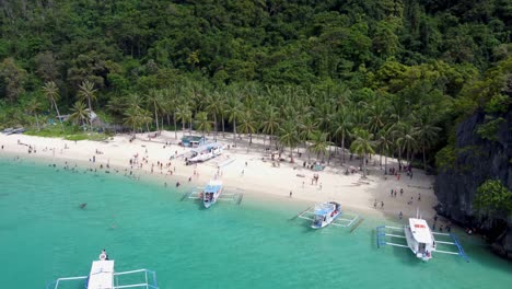 Vista-Aérea:-Barcos-De-Banca-Filipinos-De-Estilo-Catamarán-Que-Atraviesan-En-Primer-Plano-La-Playa-Tropical-De-Siete-Comandos,-Turistas-Nadando-En-Aguas-Turquesas-Claras-Sobre-Arena-Blanca