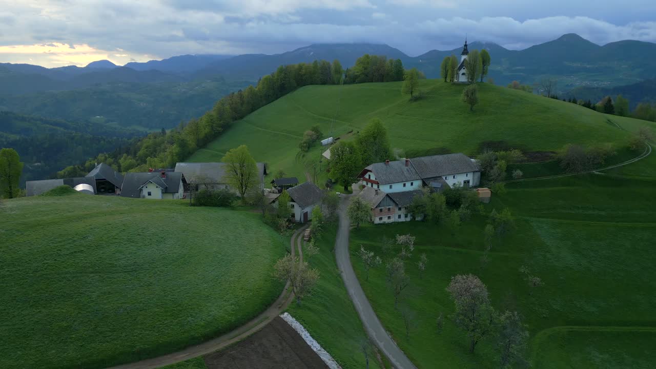 Auringonlaskunäkymä viehättävästä Surujen Neitsyt Marian kirkosta Bukov Vrissä, Gorenja Vas–Poljanessa, Sloveniassa – Kiertävä dronekuva