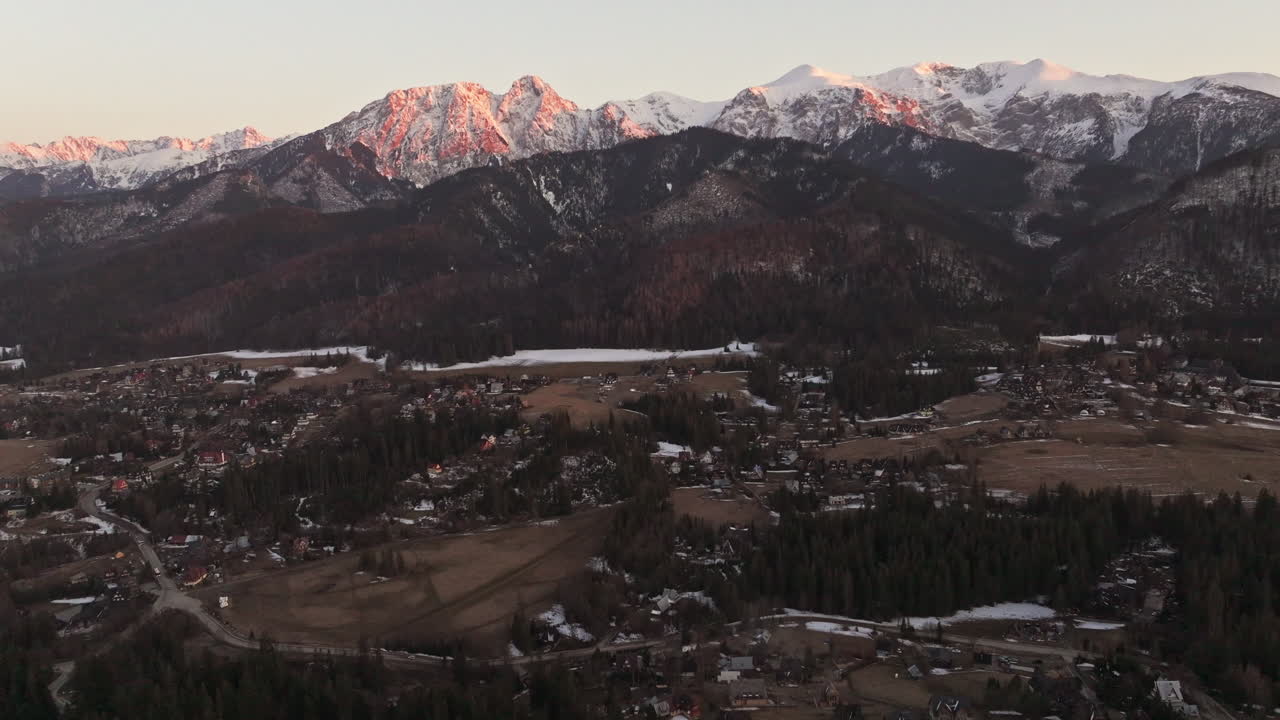 Rugged terrain stretches toward distant horizon under a bright clear sky, aerial tilt up from mountain town to ridges