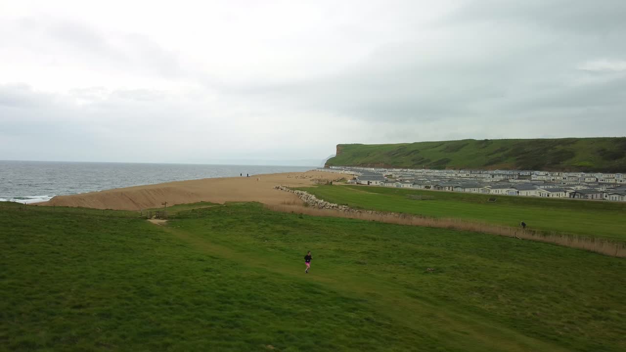 Aerial footage rotates around a female running across a field next the to a south coast beach. The Jurassic coast, Dorset, the female is running away inland as the footage rotates to the right.