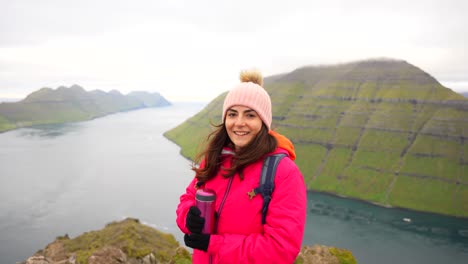 woman smiles happily while admiring view from klakkur mountain peak holding a thermos, faroe islands