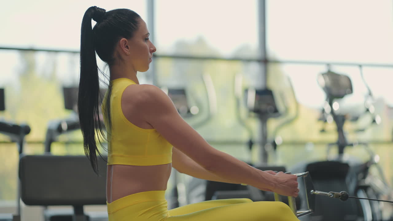 Hispanic woman does exercises on the rowing machine. A girl sits behind a simulator is called a rowing machine. sitting pulls the weight of large Windows and treadmills of the gym