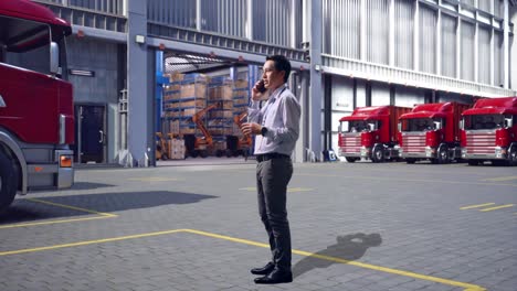 full body side view of an asian male professional worker standing, outside of logistics distributions warehouse, he is speaking with someone in the phone and moving his left hand