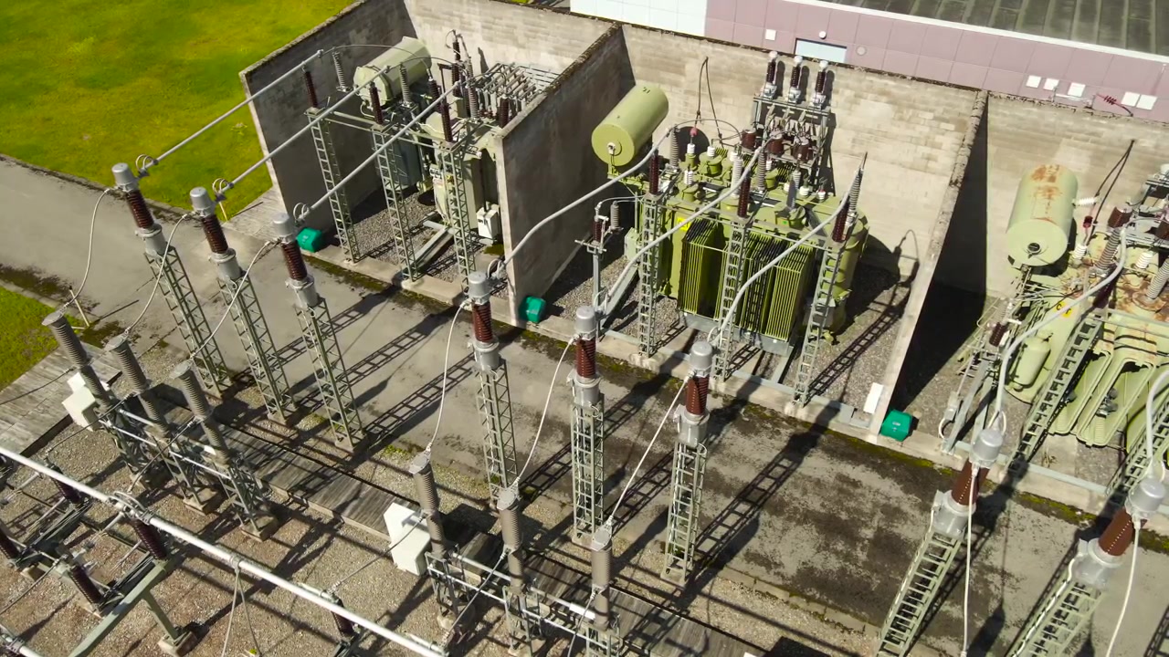 Letecký pohled shora na vysokonapěťovou elektrickou rozvodnu s transformátory, izolátory a elektrickým vedením, ukazující infrastrukturu pro výrobu a distribuci elektřiny.