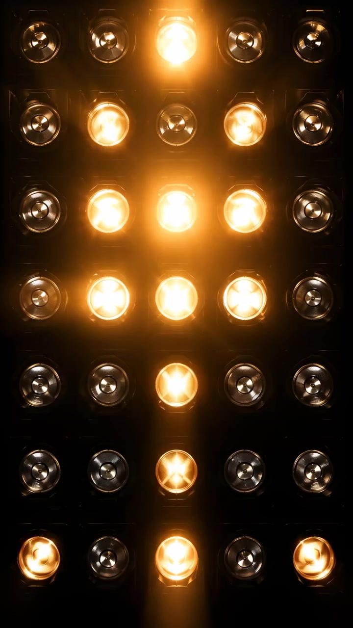 A centered grid of warm amber spotlights shines in a dark setting, with several lamps lit and others fading into shadow. The strong contrast and symmetrical