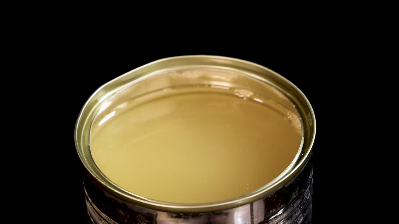 Cooked green peas fall into an open can filled with liquid, creating splashes and bubbles. Studio lighting highlights the metallic can against a black background