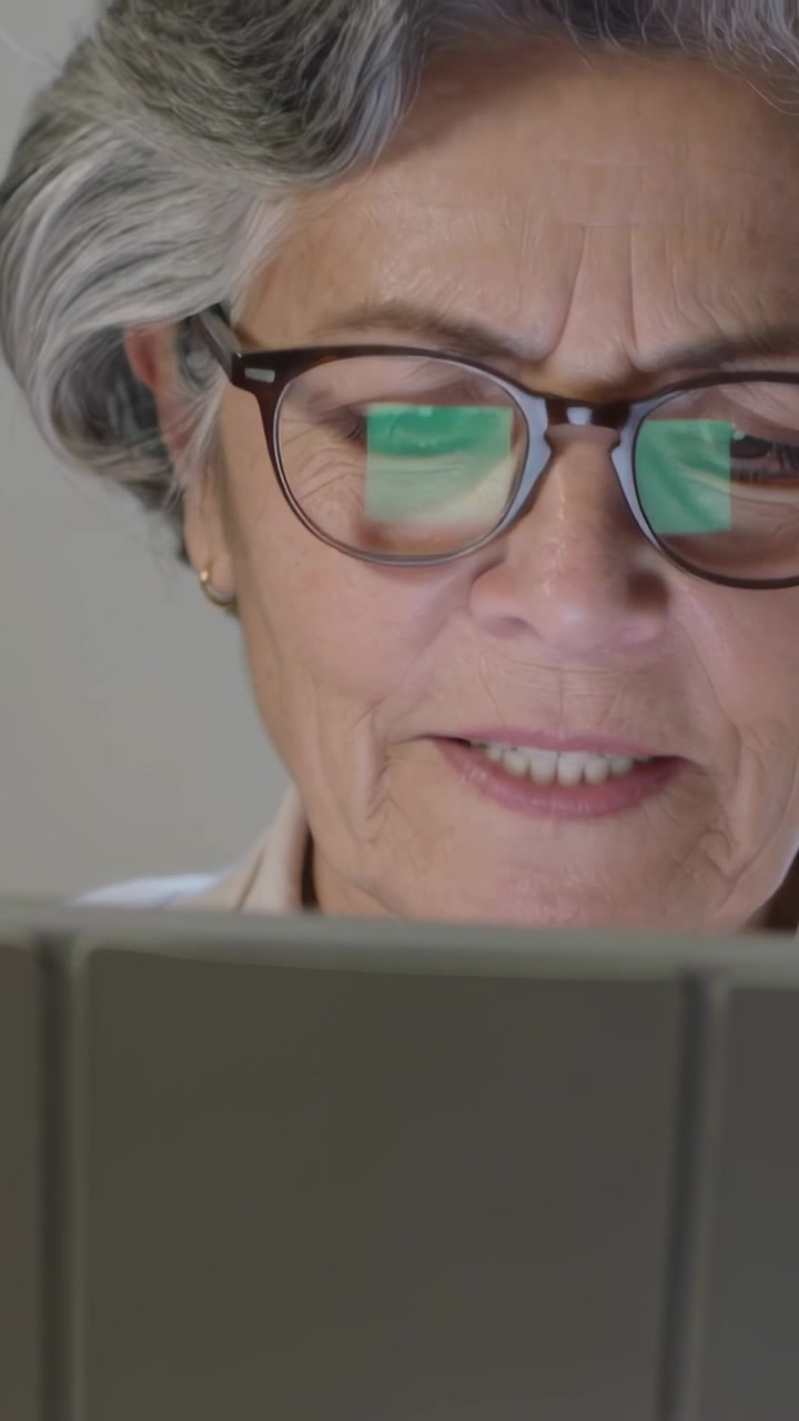 Video verticale: Anziana che legge e controlla un tablet in una stanza, mormorando e poi sorridendo con lenti verdi.