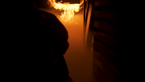 firefighter walking down narrow hall toward roaring hot inferno kneels down and begins spraying water from fire hose toward the flames to extinguish them