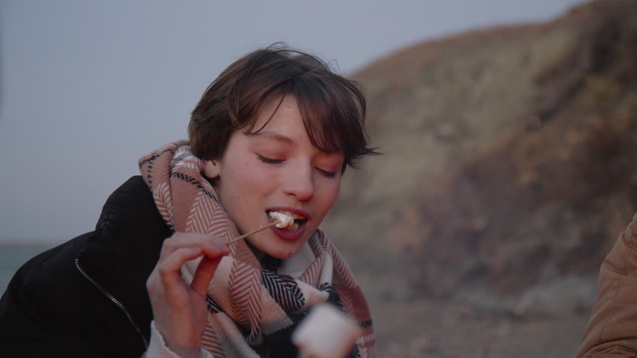 카메라는 친구와 함께 모닥불 주위에 앉아 마시멜로를 굽고 먹는 십대 소녀에게 초점을 맞춥니다.