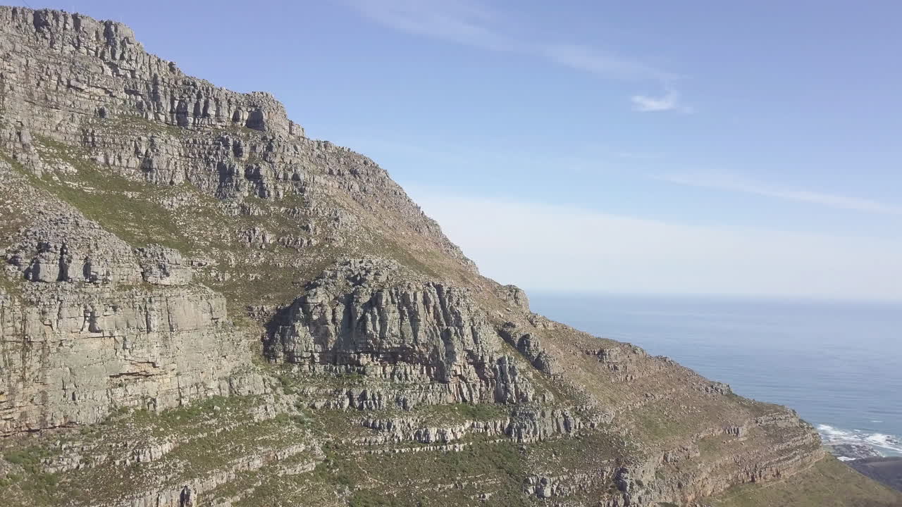 Suggestiva vista aerea con drone in ascesa sopra Table Mountain, con le sue ripide e maestose scogliere rocciose di arenaria, sullo sfondo di un luminoso e pittoresco Oceano Atlantico blu e di un cielo con nuvole cirri.
