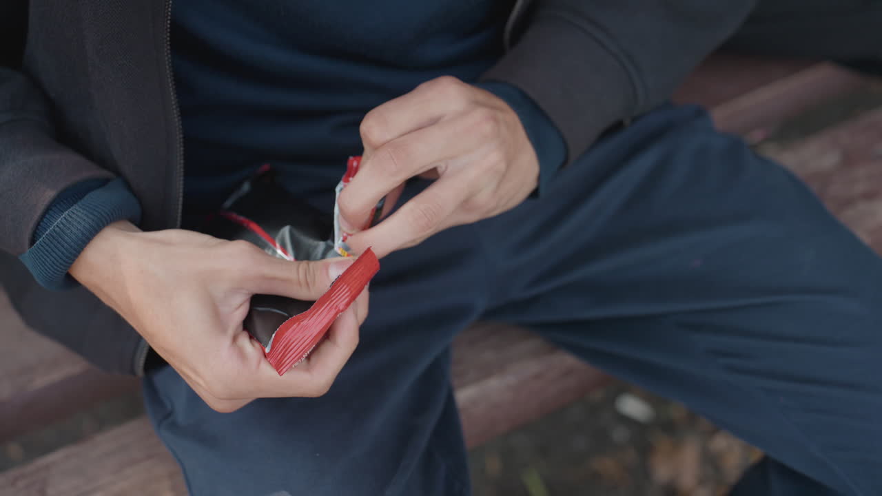 mani che scartano un pacchetto di oggetti di scena rosso su una panchina, primo piano con dita concentrate che preparano un pacchetto lucido durante le prove in strada, giacca casual e pantaloni blu navy visibili, gesti tesi e deliberati e spontanei