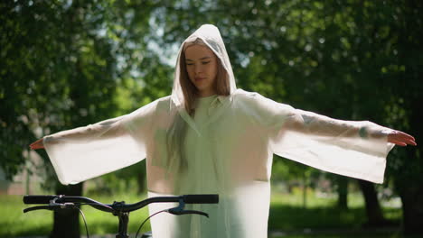 young woman in translucent raincoat stands with arms stretched out, gently swaying her hands close to her bicycle on sunny day, surrounded by lush greenery