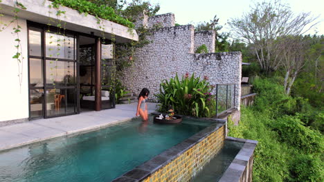 Young-Asian-Woman-in-Swimsuit-Enters-Infinity-Swimming-Pool-with-Floating-Breakfast-on-Tray-in-a-Luxury-Bali-Villa-at-Sunrise,-Aerial-Wide