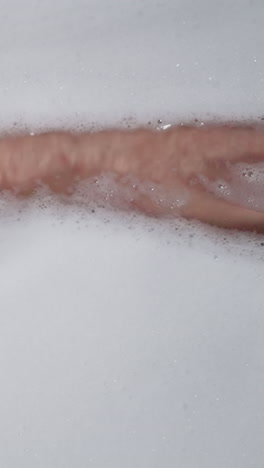 woman leg hides under soapy foam in bathtub closeup. lady washes body taking antistress bath with cosmetic products at home. woman enjoys body care routine