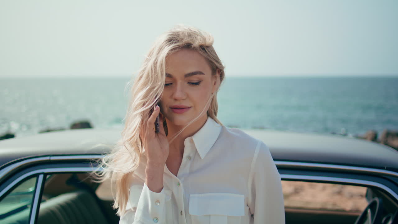 레트로 자동차 근처의 아름다운 해변에 서 있는 스마트폰으로 말하는 금발의 초상화.