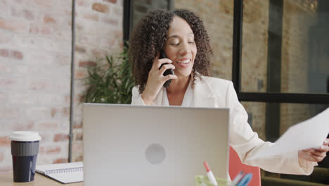 happy biracial businesswoman at desk with laptop holding document and talking on phone, slow motion