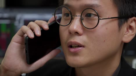 young asian businessman in glasses engaged in a phone conversation with blurred stock market screens in background, close-up