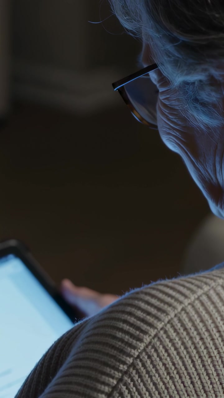 Vertikální video: Tablet osvětluje starší ženu čtoucí tablet v přítmí místnosti, s brýlemi a svetrem, s volným místem pro text.