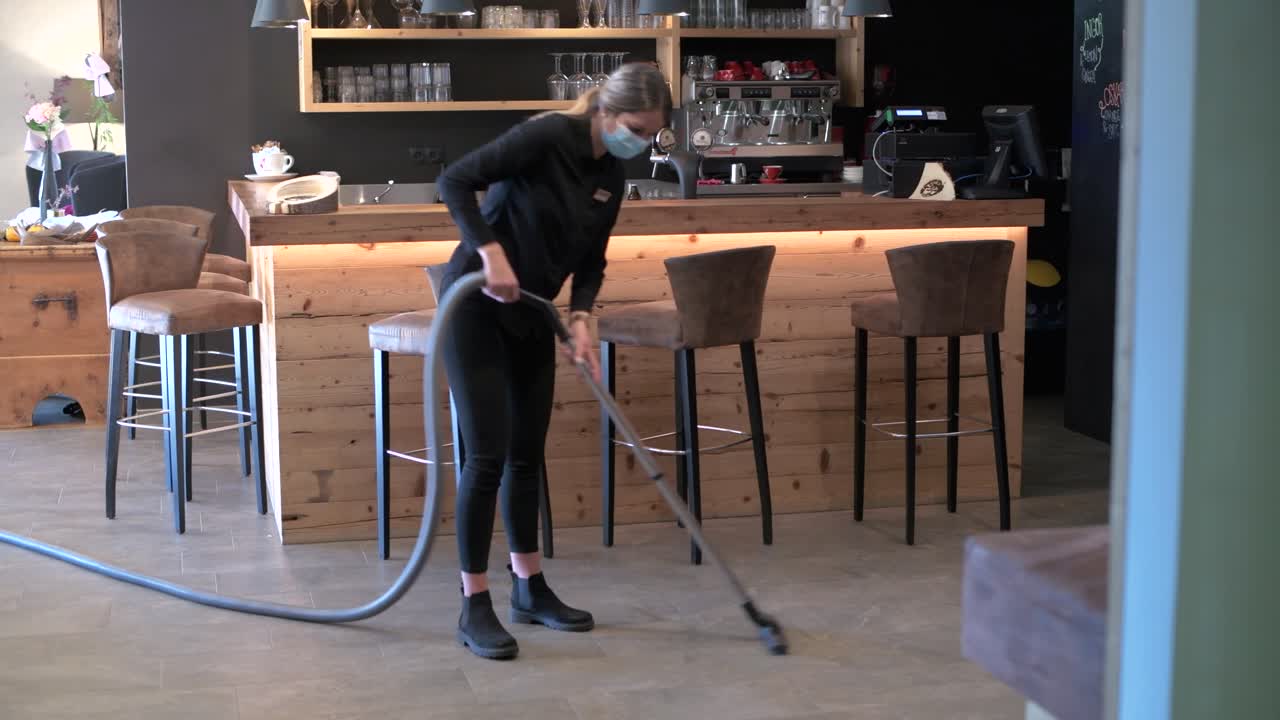 안면 마스크를 쓴 웨이트리스가 레스토랑과 바 지역을 진공 청소기로 청소하고 있습니다.