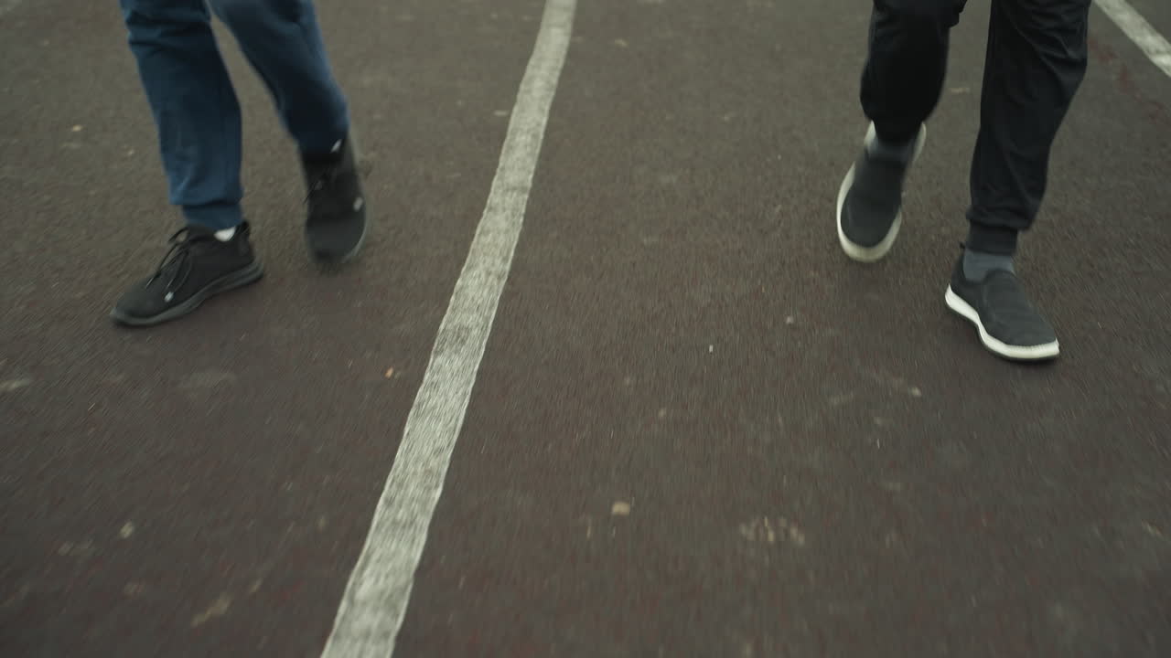 Close up leg view of two individuals wearing joggers and sneakers, walking on a track near a grassy field before transitioning into a jogging pace