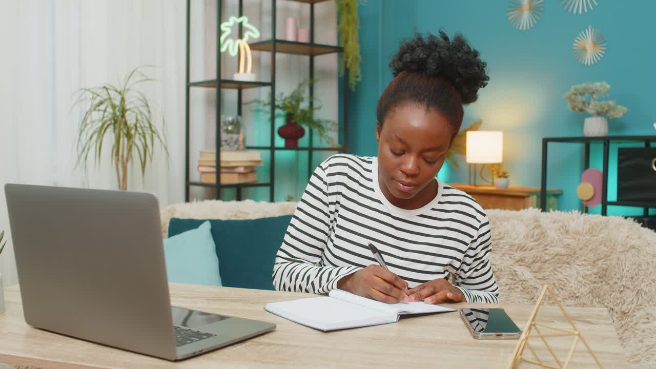 African american woman working focused on laptop sitting home on sofa writing notes in living room