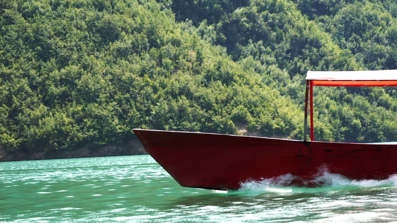 Albania, Lake Koman, prow of a red boat on the lake's surface, with a wooded shore in the background