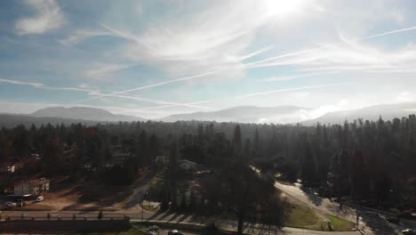 aerial flyover small forested mountain community, mountains in background, big sky with clouds and sunlight breakthrough