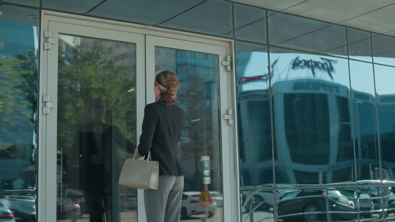 Office worker walks into office building carrying handbag, dressed professionally in black jacket and gray trousers, with glass door reflecting urban street, parked cars, and cityscape on sunny day