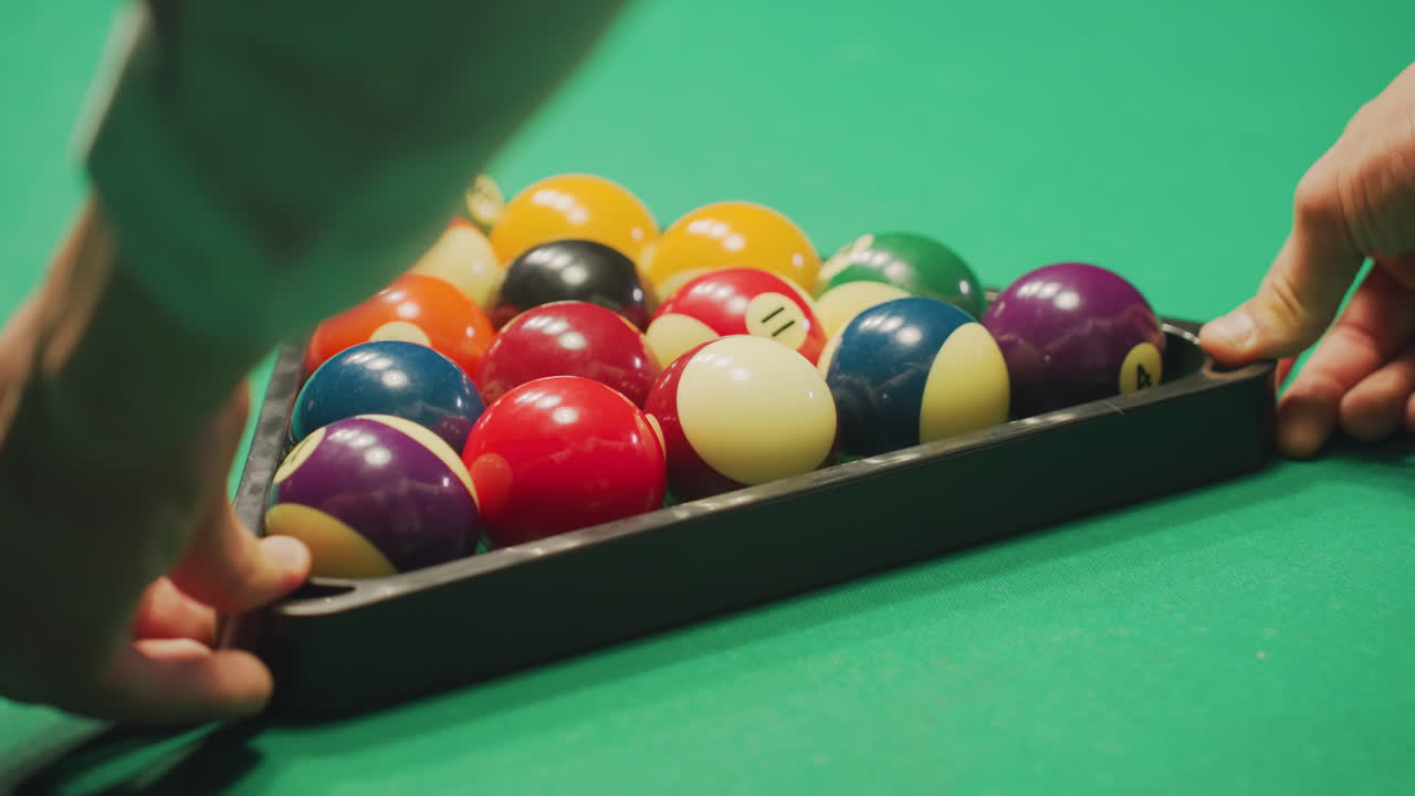 Close-up back shot of young hand in white shirt gently rolling a well-arranged set of colorful billiard balls in triangle formation on green pool table
