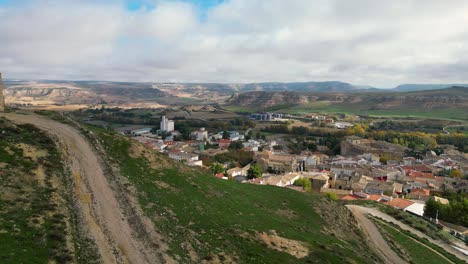 Vuelo-Sobre-Una-Colina-Con-Restos-De-Una-Fortaleza-Medieval-Descubriendo-Un-Pueblo-Con-Un-Antiguo-Palacio-Y-Campos-De-Cultivo-Con-Montañas-Bordeadas-De-Un-Cielo-Con-Nubes-En-Una-Mañana-De-Invierno-Cuenca-España