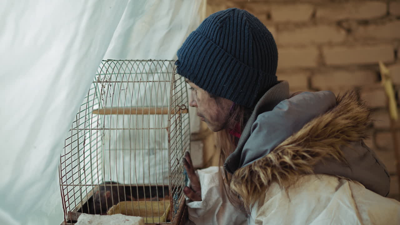 Person with dirty face wearing winter jacket and hat leans forward to closely observe rat inside rusted cage placed near cold brick wall, expressing curiosity in abandoned space