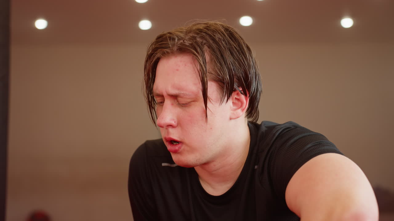 Male fighter exhausted after intense workout in gym, breathing heavily, face red with fatigue, wearing black sportswear, showing physical strain, effort under bright training lights