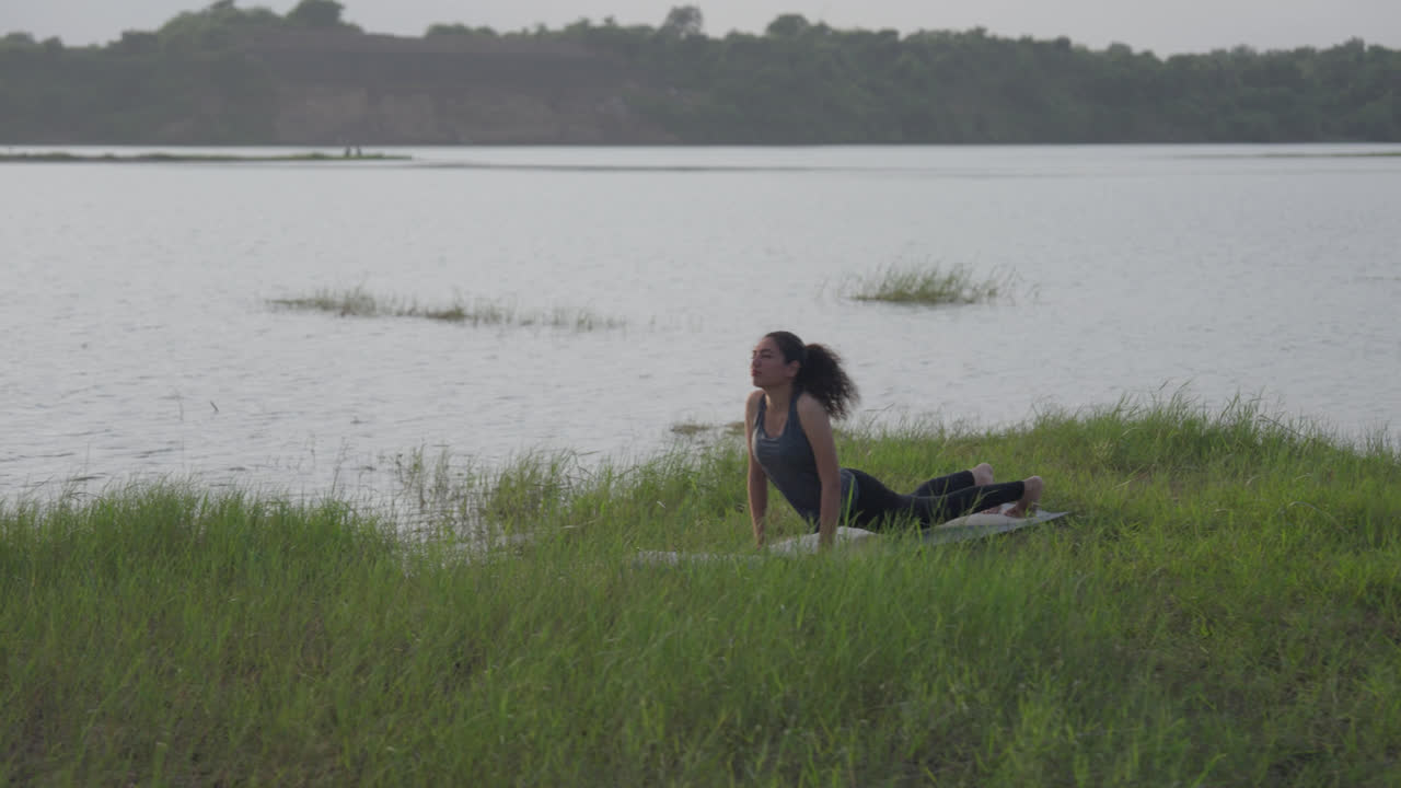 A slow-motion of South Asian Woman in Upward-Facing Dog (Urdhva Mukha Svanasana) doing yoga by a lake in tall grass